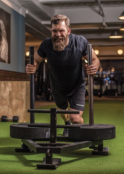 GaylenK pushing a sled of weights across a grass floor in a gym